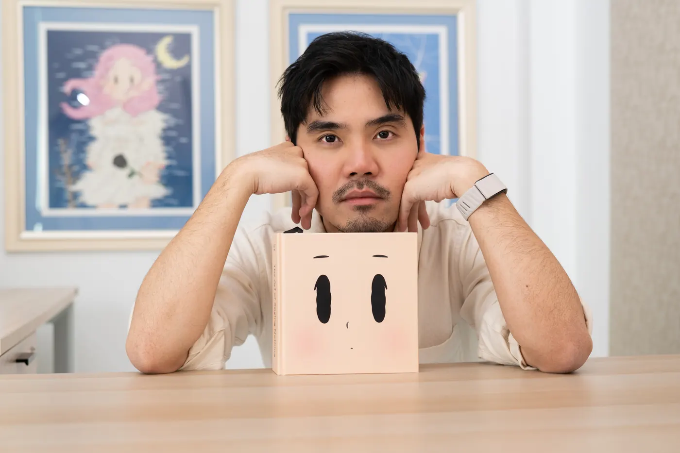 Tum Ulit resting his chin on his hands behind the book, framed prints of his work behind him